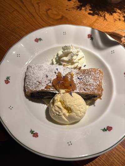 A slice of strudel dusted with powdered sugar sits on a decorative plate alongside a dollop of whipped cream and a scoop of vanilla ice cream. A spoonful of what appears to be apple compote and a few small, dried flowers are on top of the strudel. The plate is decorated with small flowers and dots around the rim and is placed on a wooden surface.
