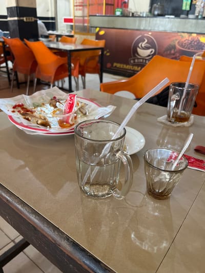 An overhead, eye-level shot shows a messy table at a restaurant in Kuta Blang, Lhokseumawe, Indonesia. The table is cluttered with used dishes and glasses. In the foreground, a clear glass mug with a handle contains a clear liquid and a white plastic straw. To its right, a smaller clear glass cup holds a spoon and a red packet. In the middle ground, a white paper plate is strewn with remnants of food, including chicken bones, and a small dish of sauce, with a red packet sticking out of it. Behind this, a clear glass with amber liquid and a straw sits on a white coaster. The background reveals orange chairs and tables, with a large brown advertisement featuring a coffee cup and coffee beans. The lighting is bright and fluorescent, typical of a restaurant environment.