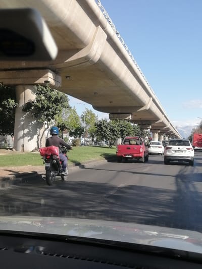 A road scene with a concrete elevated railway overhead. A motorcyclist wearing a helmet with a red delivery pack is visible on the left lane. A red pickup truck, a white sedan, and a white SUV are in the adjacent lane. Greenery divides the lanes. The image appears to be taken from the dashboard of a vehicle. Mountains are visible in the distance under a clear blue sky.