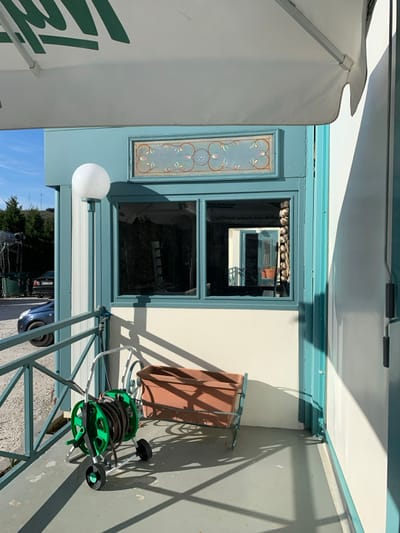An outdoor scene in Kastraki, Greece, captured on a bright, sunny day, shows a section of a building with a patio or veranda. The sunlight casts strong, defined shadows across the concrete ground and white walls, indicating midday or early afternoon.
A prominent feature is a window with a blue-green frame, set into a light-colored, possibly cream or off-white, wall. To the left of the window, a decorative, elongated glass panel features intricate floral patterns in light blues, pinks, and greens. Above the window, what appears to be a light-colored awning or canopy stretches across, partially obscuring the upper part of the frame. On the far left, a white umbrella or awning shows green lettering.
Through the window, a reflection reveals an infinite series of what appear to be garlic bulbs hanging in rows, alongside mirrored text that reads "ΠΕΤΡΩ... ...ΓΕΥΣΕΙΣ" (Tastes) and "...ΑΡΩΜΑΤΑ" (Aromas) in Greek, suggesting a restaurant or shop interior. Below the window, a spherical white outdoor light fixture is attached to the wall.
On the right side of the image, a brown, rectangular object, possibly a folding sun lounger or table, stands against the wall, casting a large shadow. In the foreground, a bright green hose reel on wheels, with a black hose wrapped around it, rests on the ground. A light blue metal railing or fence-like structure runs diagonally across the lower portion of the image. Through the gaps in this structure and reflected in the lower part of the window, a blue car and some distant trees are visible under a clear blue sky. No people are present, and the scene appears still.
