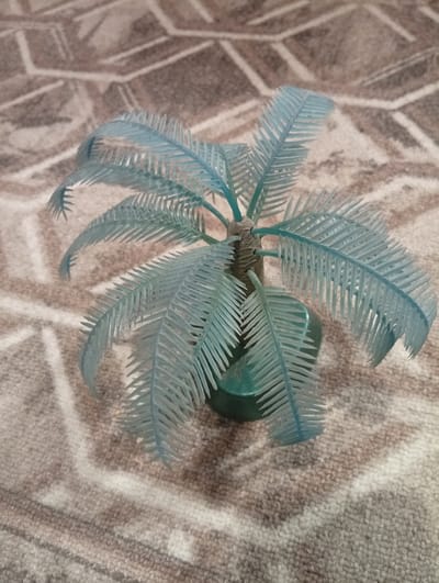 A light blue artificial palm tree stands on a brown and beige carpeted surface. The palm tree has a textured brown trunk and numerous delicate, feather-like fronds. The fronds are curved and fan outwards from the trunk. The base of the tree is a dark green plastic pot. The carpet has a pattern of light beige lines intersecting on a brown background. There are no people or animals visible in the image, and no specific location in Iskitim, Russia is identifiable.