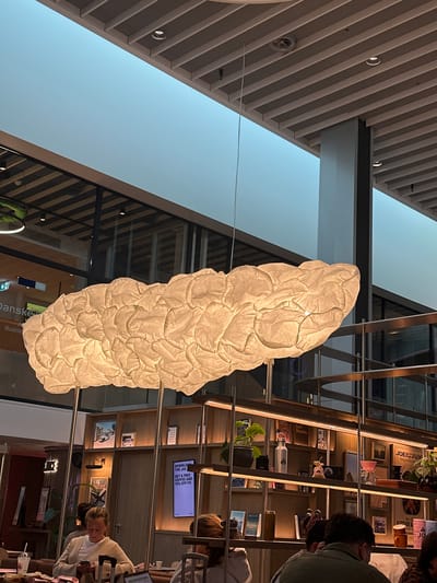 The image shows the interior of a business lounge. Overhead, a ceiling structure with recessed lights is visible. A large, cloud-shaped decorative light fixture hangs prominently in the center. To the right, there is a multi-tiered wooden shelving unit decorated with items and filled with individuals seated. A woman in a light-colored sweater is seated at the right edge of the frame. The scene is indoors and appears to be a comfortable setting in Kastrup, Denmark.