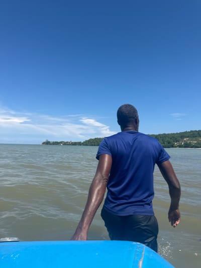 An adult male, observed from the rear, stands in brownish-green water in the coastal area of Rio Bueno, Jamaica. He has short, dark, slightly graying hair and dark skin. He wears a dark blue short-sleeved t-shirt and black shorts. His lower body, from approximately mid-thigh down, is submerged. His arms hang by his sides. In the immediate foreground, the top surface of a bright blue watercraft, possibly a boat or kayak, is visible, extending across the bottom quarter of the image, with a metallic silver cap on its left side. The water shows minor ripples. In the middle ground, a distant green coastline is visible, densely covered with vegetation and trees. Several structures are discernible on the distant land, including a building with a reddish-colored roof or spire on the left side of the landmass and various white or light-colored buildings. The sky above is clear and bright blue, with sparse, wispy white clouds near the horizon. The scene is illuminated by strong daylight.