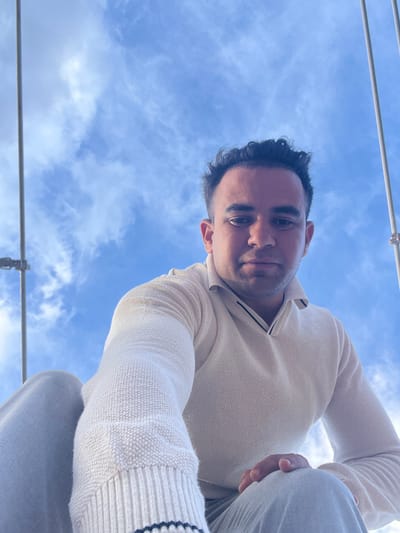 A low-angle shot shows a man looking down at the camera, with a blue sky featuring some clouds in the background. The man has dark hair and is wearing a light-colored sweater. Above and around the man is a mostly clear, light blue sky with a few clouds. The presence of metal bars or wires suggests the image might have been taken in Ait Mazigh, Morocco, on a rooftop or high place. The setting is outdoor with sunny conditions.