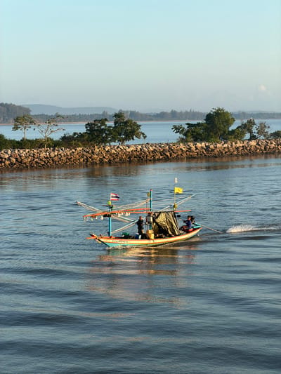 A fishing boat with two people aboard is moving from left to right across the frame on a body of water. The boat is brightly colored with a yellow and blue hull. Two flags, one with Thai colors and another yellow, are attached to a pole on the boat. A dark tarp is covering part of the boat's interior. The water is calm with visible ripples, and the wake behind the boat indicates it is moving. In the background, a rocky breakwater covered with trees separates the water from land. The sky is clear and pale blue, with distant land visible on the horizon. The location is the Pak Nam Chumphon estuary in Thailand.