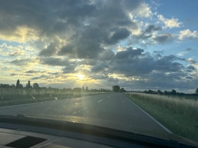 The image depicts a straight asphalt road cutting through a rural landscape in Hockenheim, Germany, under a dramatic sky. The sun is partially obscured by a mix of dark, textured clouds and patches of bright, blue sky, with visible sunbeams breaking through. To the left and right of the road, fields of tall, dry grass and green vegetation stretch towards a distant treeline. In the background, a few trees are visible, and the landscape appears flat. The perspective is from inside a vehicle, with the dashboard and windshield wipers in the foreground, suggesting the photograph was taken while driving. There are no people or animals clearly visible in the scene. The overall impression is of a serene, yet dynamic, natural environment on a cloudy day, typical of the German countryside.