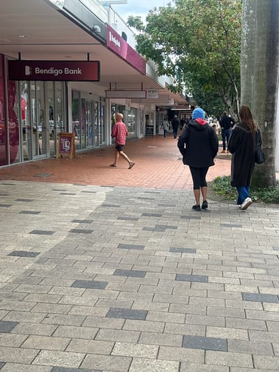 In Coffs Harbour, Australia, a person with a bald head, wearing a red and black checkered shirt and black shorts, walks on a paved sidewalk. Behind them, two women walk away from the camera. The woman in front wears a black jacket, black leggings, and a blue beanie. The woman behind her wears a dark brown coat and blue pants. The sidewalk is paved with light gray and dark gray bricks. Shops line the street, including a Bendigo Bank with a prominent sign. Trees and a concrete pillar are visible on the right side of the image. The sky is overcast.