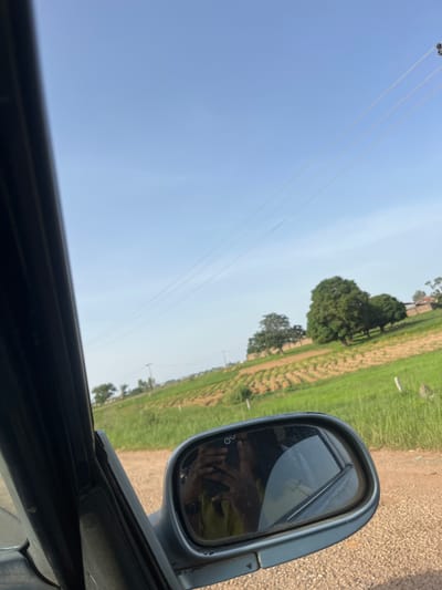The image depicts a view from inside a vehicle, looking out towards a rural landscape. A side-view mirror is prominently featured, reflecting the image of a person holding a phone, presumably taking the photo, and the interior of the car. Outside the vehicle, a dirt road leads to a field with rows of crops, likely agricultural land. Several trees are scattered across the landscape, and in the distance, utility poles and power lines are visible against a clear blue sky. The setting appears to be a sunny day in a rural area of Ladura Foron, Nigeria.