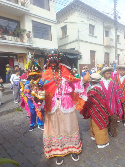 This image depicts a vibrant street scene in Amaguaña, Ecuador, featuring a cultural procession. In the immediate foreground, a person wears a large black ceremonial mask with red lips, white eyes, and an open-mouthed expression, adorned with numerous multi-colored beaded necklaces. This figure is dressed in elaborate traditional attire consisting of a bright red shawl with orange fringe and an embroidered blue bird, layered over a pink satin-like blouse with white lace, and a long, light beige skirt featuring intricate colorful embroidered bands at the hem. Black and white sneakers are visible on their feet.
To the left and slightly behind, a second, shorter individual, also masked in black with a more adorned facial mask and a tall, conical headdress embellished with colorful beads and fabrics, wears a blue and gold satin-like ensemble with orange fringe and a short, multicolored poncho. They wear black sneakers.
To the right of the central figures, two boys are dressed in traditional Ecuadorian attire, including light-colored fedora-style hats, striped ponchos (one red and pink, the other red and black), and shaggy, fur-like brown trousers or leg coverings. One boy holds a light brown, gourd-shaped object.
The scene unfolds on a dark grey cobblestone street where small patches of green grass are visible between the stones. In the background, light-colored, multi-story buildings line the street. The building on the left features a balcony with potted plants, including red flowering plants, and a ground-level shop entrance. The building on the right includes multiple windows and a small concrete balcony, with a dark utility pole and numerous overhead wires present against a partly cloudy sky. Numerous other adults are visible in the background, some observing the procession from the sidewalks.
