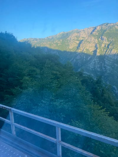 A high-angle, wide shot captures a sunlit mountain range and lush green forested slopes under a clear blue sky. In the foreground, a light-colored metal railing with two horizontal bars runs diagonally across the bottom of the frame, suggesting a viewpoint from a bridge or elevated walkway. The railing appears weathered and slightly rusted in places. The forested slopes descend from the left of the frame, dappled with shadows. The rugged, rocky peaks of the mountain range dominate the upper right portion of the image, with steep, textured cliffs. A prominent diagonal shadow cuts across the mountain and the upper part of the forest, indicating the sun's position and the time of day, likely late afternoon. The scene is natural and devoid of human presence. The location is described as being in the city of Sela, Montenegro.
