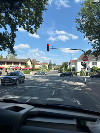 The image captures a street scene in Giessen, Germany, on a sunny day with scattered clouds. The perspective is from inside a vehicle, looking out. The road is paved and marked with white lines, including a bicycle lane. Several cars are visible on the road. A traffic light is illuminated with a red signal, indicating that traffic should stop. A prominent "STOP" sign is also visible next to the traffic light. In the background, there are buildings, some with awnings, and lush green trees lining the street. The overall impression is that of a typical urban or suburban environment on a clear day, with traffic at a standstill due to the red light. No people are explicitly visible in the scene. The license plate of the black car in the foreground reads "GI-NE 1222". The name "La Perla" is visible on the awning of one of the buildings.
