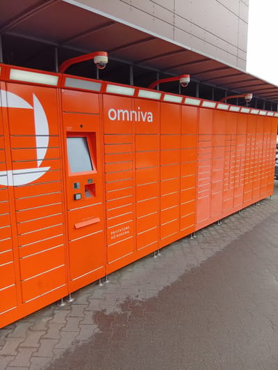 The image shows an outdoor Omniva parcel locker terminal, which is bright orange and features a touchscreen interface and multiple locker compartments. Two security cameras are mounted on orange brackets above the lockers. The terminal is situated on a paved area with cobblestone-like patterns, and there are puddles of water on the ground, suggesting recent rain. In the background, a modern building with a light-colored facade is visible. The text "omniva" is prominently displayed on the terminal, along with some Lithuanian text that appears to be service information. There are no people visible in the image. The scene is consistent with a public amenity in an urban or suburban setting, and the presence of the Lithuanian text suggests it is located in Lithuania. Specifically, this Omniva terminal is located in Kuršėnai, Lithuania.