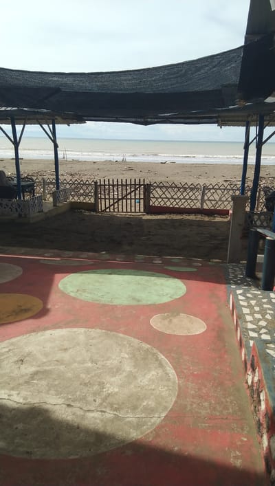The image depicts a view of the beach in Reudeuep, Indonesia, as seen from a shaded outdoor seating area. The foreground features a red concrete floor decorated with large, irregularly shaped circles in grey and light brown, as well as smaller light brown circles. To the right, a low wall with a mosaic of white and blue tiles is visible. Beyond this, the ground transitions to a sandy area enclosed by a white fence with a lattice pattern and a wooden gate. Further back, the beach stretches towards the sea, with gentle waves lapping the shore. In the background, the horizon is marked by the ocean meeting a pale, overcast sky. A dark, mesh-like canopy supported by blue poles provides shade over the foreground and a portion of the sandy area. There are no people visible in the scene.