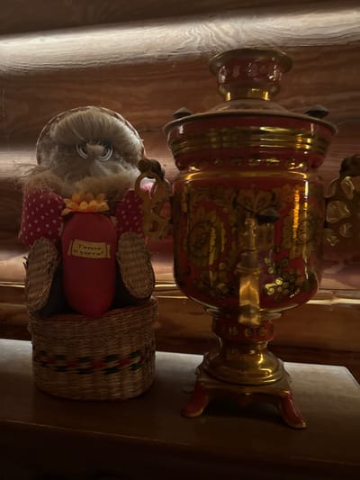 The image depicts a still life composition featuring a plush toy and a samovar, placed on a wooden surface against a background of rough-hewn wooden planks. The plush toy, positioned to the left, is light brown and white with large blue eyes and a fluffy mane. It is wearing a red and white polka dot fabric over its shoulders. Its body is a red, heart-shaped object with a yellow flower on top and a small banner that reads "Тепла и уюта!" in yellow lettering. The toy is seated in a woven basket. To the right of the toy stands a traditional Russian samovar, predominantly red with intricate gold floral patterns and ornate detailing. The samovar has a spigot and a lid. The lighting in the scene is warm and somewhat dim, casting shadows and highlighting the metallic sheen of the samovar and the textures of the wood. The location is identified as Heiligenrode, Germany.
