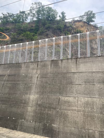 The image shows a retaining wall made of concrete planks. Above the wall, a metal mesh fence is visible, and beyond that, a rocky hillside with trees and vegetation. Overhead, power lines are strung across the scene, and the sky is overcast. There are no people or animals visible in the image. The location is identified as Dongguan, China.