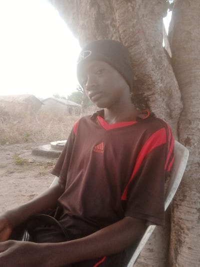 A young man wearing a dark-colored Adidas t-shirt and beanie sits in a white plastic chair. He is leaning against a large, textured tree trunk. In the background, there is dry, sparse vegetation and a few buildings. The light is bright, and the scene has a rustic, outdoor feel. The man is the central subject, drawing the viewer's attention with his calm expression.