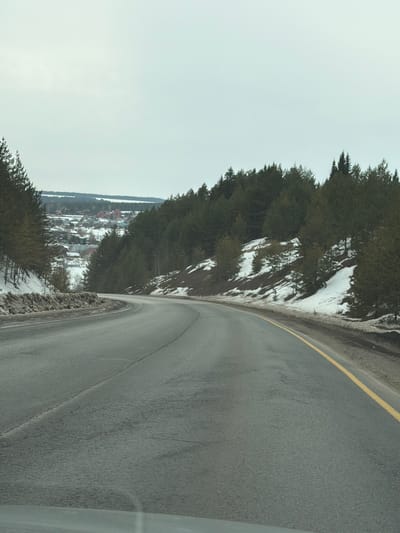 The image captures a two-lane asphalt road curving to the left, likely observed from a vehicle in the vicinity of Doksha, Russia. The dark gray road surface exhibits some dampness. A solid yellow line delineates the right edge of the pavement, and a faint white broken line is visible near the center. On both sides, snow-covered embankments ascend, populated by dense evergreen trees and some bare deciduous trees. Patches of exposed ground and sparse vegetation are visible where snow has melted. In the mid-distance, a village is visible in a valley to the left of the road, consisting of multiple houses with predominantly red, dark, and light-colored roofs. Beyond the village, a body of water is discernible, bordered by a distant treeline. The sky is uniformly overcast and light gray. No individuals or other vehicles are present.