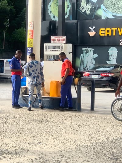 This image depicts a daylight scene at a fuel station in Lakowe, Nigeria. Three adult males are present near a white fuel pump. On the left, a male in a blue uniform with red accents is observed looking down at an object held in his hands. In the central foreground, a male wearing a light-colored suit with a distinct blue floral pattern stands facing a fuel pump. To the right, a male in an orange shirt and blue trousers is actively dispensing fuel from the pump nozzle into a large, yellow plastic jerrycan positioned on the ground.

The fuel pump features a digital display and has a prominent red sign above it stating "COMPLAINTS? 0817 206 8427 ics@11plc.com". Adjacent to the pump, a light-colored pillar displays various warning signs, including "NO SMOKING," "TURN OFF PHONES," "DANGEROUS AREA," and "TURN OFF ENGINE." In the background, a black Toyota Camry sedan with the Nigerian license plate "7974DCA" is parked. Behind the vehicle and pump, a commercial building with a dark, textured facade is visible, featuring large graphic designs and a prominent orange sign displaying "EAT WITH HIME" alongside a bowl and spoon logo. The ground surface is a textured, light-colored pavement. A partial view of a bicycle or motorcycle wheel is visible on the far right of the frame.