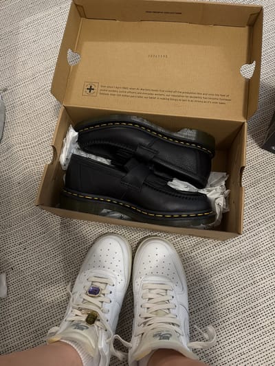 The image presents an indoor scene, likely in a home in Hainburg an der Donau, Austria, showcasing a pair of new black Dr. Martens loafers inside their cardboard box, positioned on a light-colored, textured carpet. Alongside the box, a person's lower legs and feet are visible, wearing white Nike Air Force 1 sneakers.
The open Dr. Martens shoebox, made of brown cardboard, contains the black loafers. They feature a textured leather finish, traditional yellow stitching along the sole, and a tassel detail on the vamp. Inside the box, the shoes are partially wrapped in white tissue paper. Printed text on the box includes "VNGX DM23#144 329X193X126MM" and "20241113". A longer text block reads: "Ever since 1 April 1960, when Dr. Martens boots first rolled off the production line and onto the feet of postal workers, police officers and everyday workers, our reputation for durability has become footwear folklore. Over 100 million pairs later, our belief in making things to last is as strong as it's ever been." The Dr. Martens logo is also visible.
The white Nike Air Force 1 sneakers worn on the feet display the "NIKE AIR" branding on the side and feature small perforations on the toe box. Each sneaker is accessorized with a prominent, rectangular, jewel-like lace charm; one is a deep blue and the other a yellow/gold, set with smaller clear stones. The overall activity depicted suggests the unboxing or presentation of the new Dr. Martens loafers, contrasted with the worn but stylized Nike sneakers. The lighting indicates an indoor setting, with no specific time of day or weather discernible.