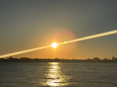 This image depicts a waterfront scene captured from Jersey City, United States, at either sunrise or sunset. The sun, a brilliant white-yellow orb, is positioned low on the horizon, slightly to the left of the frame's center. It generates a prominent, strong diagonal lens flare extending from the bottom-left to the top-right, along with a circular orange-red halo effect around its core. The sky exhibits a gradient, transitioning from a warm golden-orange near the horizon to a muted blue-grey at the upper edge. A dark, silhouetted landmass stretches across the entire horizon, composed of a mix of trees and low-rise buildings, including at least one structure with a distinct dome or tower visible on the right side. The water, which occupies the lower half of the frame, is a dark blue-grey. It reflects the sun's light as a shimmering, golden path extending from the horizon towards the foreground. Small ripples are visible on the water's surface. No human subjects or discernible activities are present within the scene.
