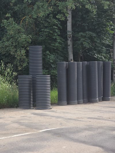 In the city of Deulino-3, Russia, a group of black, corrugated plastic pipes are arranged on a dirt and asphalt surface. The pipes are of varying heights, with the tallest standing significantly higher than the others. They are positioned in front of a dense backdrop of green trees and foliage. A slender, pale tree trunk is visible to the left of the pipes, partially obscured by the vegetation. The ground in the foreground is a mixture of dirt and asphalt, with a white line marking a boundary on the asphalt surface. The scene appears to be outdoors, possibly in a storage or construction area.