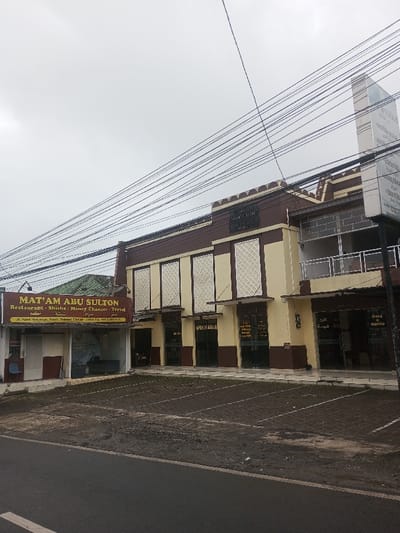 The image displays a street-level view of a commercial building complex under an overcast sky in Palasari, Cipanas, Bogor, West Java, Indonesia, located at Jl. Raya Puncak. The main structure is a two-story building with a brown lower facade and cream-colored upper sections featuring white decorative panels. Multiple ground-floor entrances with glass doors and brown awnings are visible. Signage includes text in Arabic and English, with one sign clearly stating "OPEN 24 JAM" (Open 24 Hours). On the far left, a single-story building with a green roof abuts the main complex. This building features a red and yellow banner sign for "MAT'AM ABU SULTON," advertising "Restaurant - Shisha - Money Changer - Travel," and provides the address: "Jl. Raya Puncak, Ps. Palasari, Cipanas - Bogor, Jawa Barat, Indonesia." In front of the buildings, an unpaved dirt and gravel parking area with faded white painted lines separates the structures from an asphalt road. A tall, narrow white vertical sign is positioned on the right side of the main building. Numerous black utility wires crisscross the upper portion of the frame against the grey sky. No individuals or vehicles are present in the scene.