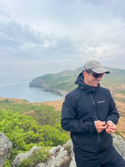 A medium shot depicts a man standing on a rocky precipice overlooking a bay. The man is clad in a black jacket, a grey cap, and dark sunglasses. He is smiling and looking to his left. His hands are raised near his chest. The jacket has the word "FINNTRAIL" on the upper left chest. In the background, a body of water stretches towards the horizon under a cloudy sky. To the left, a green, sloping cliff extends into the water. To the right of the man, there is green vegetation growing on the rocky terrain. The location is Gvozdevo, Russia.