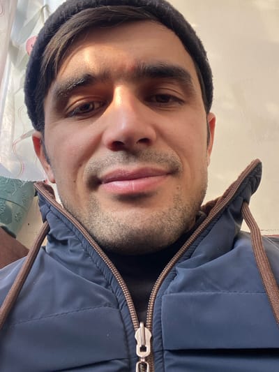 A man's face fills the frame, tilted slightly. He wears a black beanie and a dark blue puffer jacket with brown trim and a visible zipper. His skin tone is medium, and he has dark hair, dark eyes, and stubble. A warm light shines on his face, highlighting his features. The background is a mix of white wall and patterned fabric.