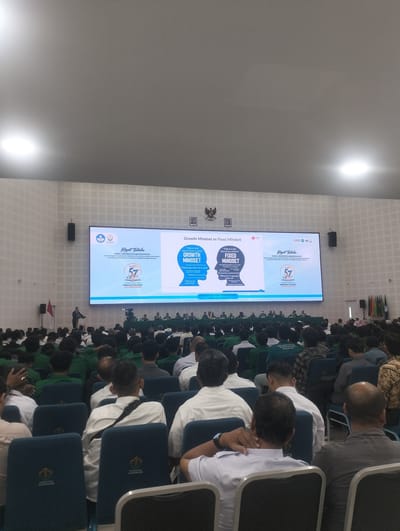The image depicts an indoor lecture hall with a large screen at the front displaying a presentation on "Growth Mindset vs Fixed Mindset". A speaker stands at a podium on the left side of the stage, addressing a seated audience. The audience members, predominantly young adults, are focused on the screen. The hall has tiered seating and appears to be moderately filled. The setting suggests an academic or professional event. The information on the screen indicates the event is held in the Auditorium, Faculty of Law, Universitas Malikussaleh, Reuleut Timu, Indonesia, on June 23, 2023.