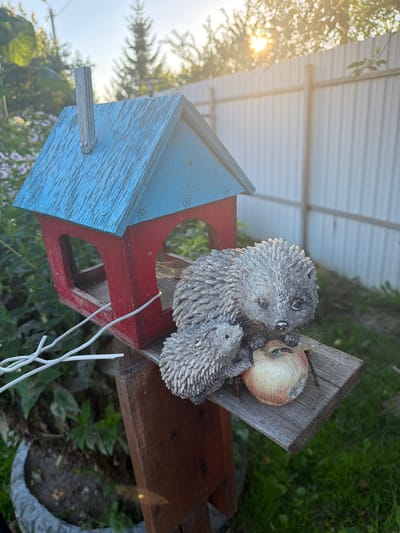 A close-up, eye-level shot captures a weathered, red and blue birdhouse in a sun-dappled outdoor setting. Two grey, textured hedgehog figurines, one larger and one smaller, rest on a wooden perch attached to the birdhouse. A single, slightly bruised apple, tinged with red and yellow, sits in front of the hedgehogs, with small twigs protruding from it. The birdhouse's blue roof shows signs of wear, and a small wooden chimney protrudes from its peak. A thin white wire snakes across the front of the birdhouse. The background features a white metal fence, lush green grass, and blurred foliage with sunlight filtering through. The scene is devoid of human presence. The image appears to be taken in the city of Primernoe, Russia, suggesting a residential or garden environment.