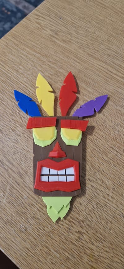 This media file depicts a brightly colored, 3D-printed object resembling a stylized tiki mask. The mask has a brown, wood-like base with several prominent features. On the left side, there are five layered, feather-like protrusions in vibrant colors: purple, red, yellow, yellow, and blue, from top to bottom. The mask itself features a central red, jagged frame with a white, grid-like interior that appears to represent a mouth with teeth. Above the mouth-like feature, there are two pairs of light green and yellow rectangular shapes, possibly representing eyes or decorative elements. To the right of the central structure, a light green, torn-edged shape protrudes, possibly representing a leaf or additional decoration. The object is positioned on a wooden surface with a distinct grain pattern. The lighting suggests an indoor setting, possibly under artificial light, as there are no strong shadows indicative of natural sunlight. No people or text are visible in the image. The object's design and vibrant colors suggest it might be a decorative item, a toy, or a component of a larger display. The setting appears to be a table or desk in a domestic or workshop environment, given the wooden surface and the nature of the object. The overall impression is one of a playful and creative handmade or 3D-printed item.