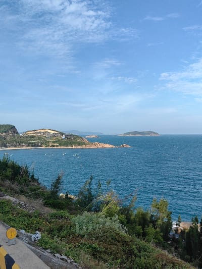 This image captures a panoramic coastal scene within Tuy Phong 4, Vietnam, under clear daylight conditions.
The foreground, specifically the bottom left quadrant, displays the edge of a concrete or paved surface, marked by a yellow and black striped bollard or barrier. Immediately beyond this, a dense layer of green and brown low-lying vegetation, scrub, and scattered small trees covers a rocky hillside sloping towards the water.
The midground is dominated by a wide expanse of deep blue ocean water, exhibiting minor surface ripples. Approximately two dozen small, light-colored boats, some with visible white buoys nearby, are distributed across a bay on the left side of the water. The coastline along this bay features a sandy beach fringed with palm trees. Ascending from the beach, a prominent green hill is visible, hosting a cluster of multi-story buildings, likely a resort or residential complex, characterized by light-colored facades and dark roofs, interspersed with vegetation. Further along this coastline, red-brown rocky headlands and smaller islets extend into the ocean.
In the background, several larger, dark green islands or distant landmasses emerge from the ocean on the right, receding towards the horizon. The sky, occupying the upper half of the image, is a clear, bright blue with scattered, wispy white clouds. No human figures are identifiable.