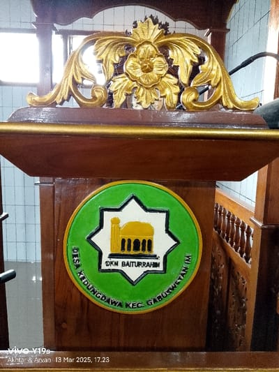 The image shows a wooden podium with a gold-colored carved floral design at the top and a circular green and gold plaque below. The plaque depicts a white eight-pointed star with a gold mosque inside and the words "DKM Baiturrahmi" inscribed on the bottom. The words "DESA KIDUNGDAWA KEC. GABUSWETAN" are inscribed around the perimeter of the plaque. The podium is positioned against a wooden wall with a carved pattern. The scene is located in the Kecamatan Gabuswetan district of Indonesia, at the coordinates -6.44 latitude and 108.028 longitude.