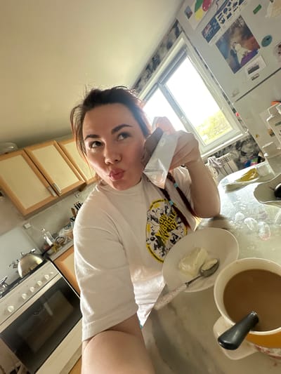 The image depicts a young woman in a kitchen. She is holding a small package, possibly of creamer, and posing for a selfie. On the counter are a mug of coffee with a spoon, and a plate with a dollop of what appears to be cream or butter. The kitchen has light-colored cabinets and an oven. A window provides natural light. There is no verifiable information to determine the location is Черёмушки, Russia.