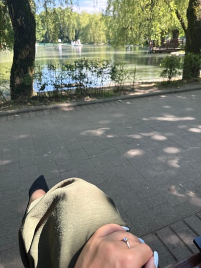 The image shows a view of a park with a lake and trees. In the foreground, a person's legs are visible, wearing a green skirt and black shoes. The person is wearing a ring on their left hand. The ground is paved with rectangular stones. In the background, a lake is visible with trees on the opposite bank. There are boats on the lake and a wooden structure on the shore. The scene suggests a sunny day in a park. The city of Khone, Georgia is not depicted in this image.