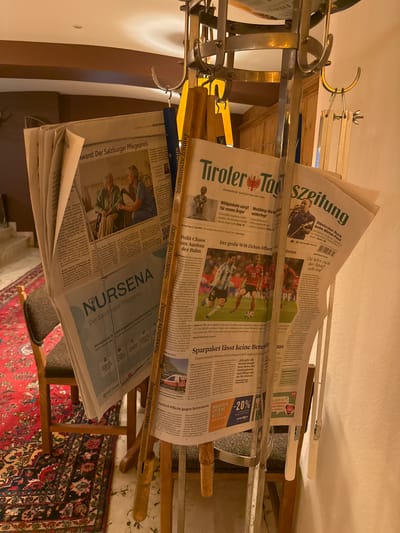 A close-up, eye-level, indoor shot focuses on a collection of newspapers held upright by a metallic stand with wooden accents. The newspapers are layered, with their headlines and some images partially visible. One newspaper in the foreground prominently displays the headline "Award: Der Salzburger Pflegepreis" and features an image of two individuals. Another newspaper to its right shows a football match in progress, with the headline "Der große WM-Zirkus öffnet". Further to the right, a newspaper with the headline "Süddeutsche Zeitung" is also visible. The backdrop consists of wooden paneling and a wall, with a red rug and a chair partially visible in the lower left. The lighting is warm and appears to be artificial. The scene is devoid of any identifiable landmarks that would pinpoint a specific location in Mayrhofen, Austria.
