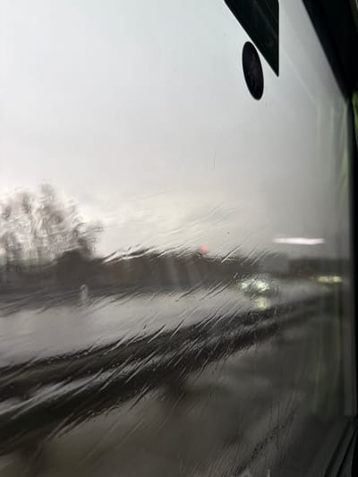The image depicts an exterior scene viewed through a rain-streaked window, likely from a moving vehicle, in Moineville, France. The foreground is dominated by the window surface, covered in numerous vertical and diagonal rivulets of water, blurring and distorting the distant view. A dark rectangular object, possibly a sticker or label, is partially visible in the upper right corner, along with a smaller circular black sticker below it.
Beyond the window, the environment appears overcast and dim. A multi-lane roadway, slick with rain, occupies the midground and lower portion of the frame. Traffic is present, with at least one vehicle displaying illuminated white headlights on the right side of the road, and another exhibiting a red taillight or brake light in the center-left. Trees or dense vegetation, their branches appearing bare, are visible on the left side of the roadway, heavily blurred by the rain and atmospheric conditions. The sky above is uniformly grey and overcast. No specific individuals are discernible.