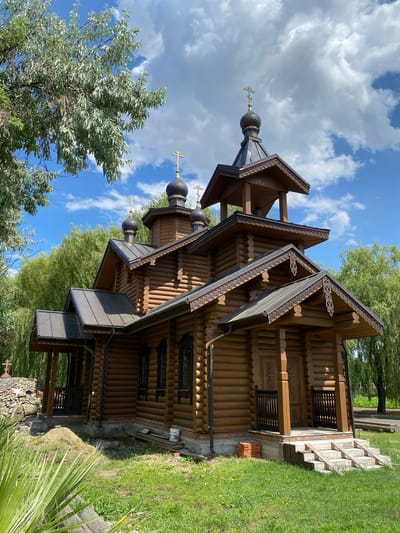 This is a vertical, eye-level, outdoor, full shot of a wooden church structure with multiple domes topped with crosses. The church is constructed from logs and features ornate trim under its layered roofs. Several windows are visible, some with decorative grates. The structure is situated amidst lush greenery, with trees and grass surrounding it. In the foreground, a palm frond is visible on the right. The sky is blue with scattered white clouds, suggesting it is daytime and the weather is pleasant. The scene appears to be a peaceful, possibly religious, setting. There is no visible text in the image. The orientation of the photo is rotated 90 degrees clockwise.