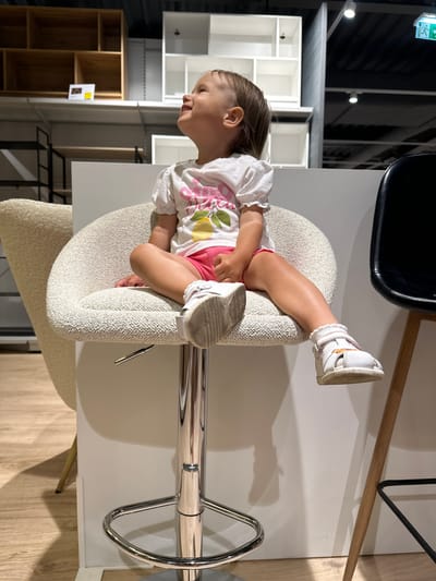 An upright, eye-level indoor shot shows a young child seated on a high, textured beige bar stool in what appears to be a modern furniture showroom or store. The child, with light brown hair pulled back, is wearing a white short-sleeved top adorned with a colorful pattern of pink flamingos (or similar birds), yellow lemons, and green leaves, along with pink shorts. They are looking downwards and to their left, with a neutral and somewhat contemplative expression. Their legs are bent, and feet, clad in white sneakers with orange accents, are resting on a part of the stool or dangling.
In front of the child is a simple white rectangular table. To the upper right, part of a black chair is visible. The background features various shelving units: white, modular open shelves on the left, and a taller, multi-compartment wooden shelving unit below them. An emergency exit sign, glowing green, is visible in the upper left corner, clearly displaying the Polish word "WYJŚCIE" (EXIT). The flooring is light wood laminate. The overall lighting is bright and even, typical of an indoor commercial space. The chrome base and footrest of the bar stool are also visible. The setting suggests a display area within a store in Ustroń, Poland.