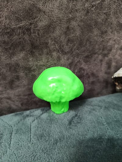 A single, bright green, plastic mushroom figurine is centrally positioned on a textured, teal-colored surface. Behind the figurine, a dark gray, plush fabric forms the background. The lighting appears to be soft, casting subtle shadows. The image does not contain any information that would allow for its location to be determined as Leninaul, Russia, or any other specific city.