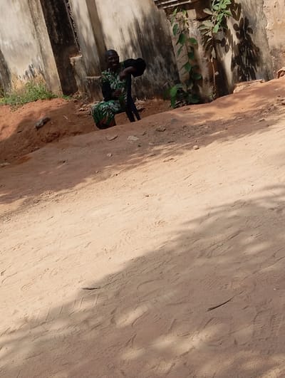 The image displays an outdoor scene in Okpatu-Ukehe, Nigeria. A man is positioned in the midground, seated or squatting behind a reddish-brown dirt mound. He has a shaved head and is wearing a patterned green and black garment over dark trousers or a skirt, smiling towards the viewer. He holds a green bottle in his right hand and a dark, possibly fabric, object in his left. A dark chair or stool is partially visible behind him. The background consists of a weathered, light-colored concrete or stucco wall exhibiting dark vertical streaks, with green vegetation growing along its right side. The foreground and midground comprise exposed reddish-brown earth, marked with tire tracks and footprints, under dappled sunlight.