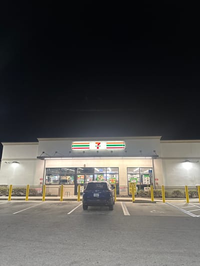 The image shows a 7-Eleven convenience store at night. A dark blue vehicle, likely a car, is parked in front of the store in a parking space. The 7-Eleven signage is prominently displayed above the entrance. The store's front is well-lit, showing product displays and windows. Parking spaces are lined with yellow bollards. The environment appears to be a typical commercial area at night in a location in the city of Homosassa Springs, United States of America.