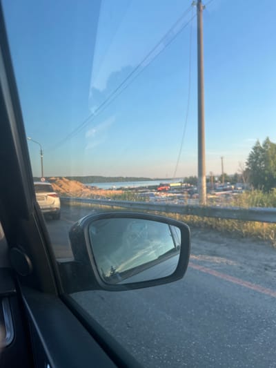The image is taken from inside a vehicle, looking out towards a roadside scene with a body of water in the background. In the foreground, the interior of the car is visible, including a side mirror which reflects the sky and trees. Outside the car, on the left, a white SUV is driving on a road with a light pole and overhead wires. To the right of the road, there is a large pile of sand, a body of water, and what appears to be an industrial or construction area with various structures and vehicles. Trees and vegetation line the right side of the road. The sky is clear and blue, indicating daytime. The location is identified as Palniki, Russia.