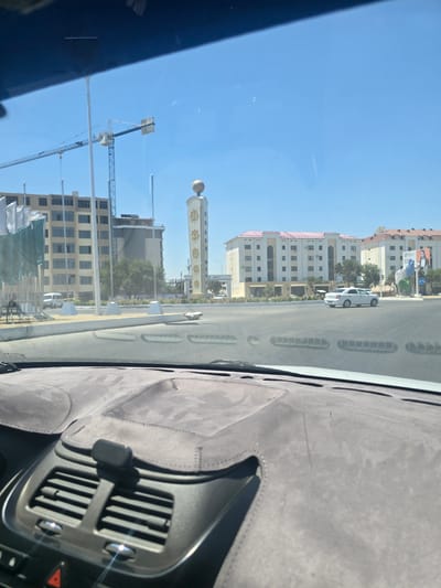 A bright, clear day is depicted through a car's windshield, looking out at a city street in Kogon City, Uzbekistan. In the foreground, the textured dashboard and air vents of a vehicle are visible. Beyond the windshield, a wide intersection is apparent, with a white sedan driving on the road. Several modern buildings line the street; one prominent structure is a tall, white tower with a golden sphere at its apex, adorned with text. Construction cranes are also visible in the background, suggesting ongoing development. Flags and trees are interspersed among the buildings, contributing to an urban landscape under a vast, blue sky.