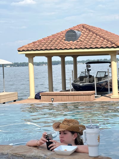 This image depicts a young woman in an infinity-edge swimming pool overlooking a lake in Montgomery, United States. The woman, approximately late teens to early twenties, with light brown hair and a woven straw cowboy hat, is partially submerged in the blue pool water, resting her forearms on the stone coping. She is holding a silver-cased smartphone with a black protective cover in both hands, viewing its screen. White nail polish is visible on her fingernails, and several rings adorn her fingers. To her left, on the pool coping, sits a white square bowl containing orange-colored snacks and a large white insulated tumbler with a handle, clear lid, and straw, featuring a yellow decal. Behind the pool, across a body of water, a large covered boat dock or boathouse is visible. This structure features a reddish-brown terracotta tile roof with a half-round louvered vent at its peak, supported by several cream-colored, fluted columns. Beneath the covered section, a dark blue and grey powerboat, equipped with a wakeboard tower, is partially visible, appearing to be elevated on a boat lift. The background reveals a distant treelined shoreline with scattered houses under a partly cloudy sky.