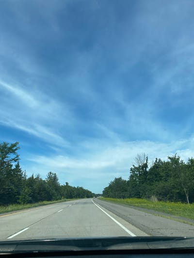 The image shows a view from the perspective of a vehicle traveling on a highway. The highway is paved and has lane markings, including a yellow line on the left and white lines separating the lanes. On either side of the highway, there are dense green trees and some yellow wildflowers. The sky is a bright blue with thin, wispy white clouds. The overall environment appears to be a rural or semi-rural setting. There are no people or other vehicles clearly visible in the immediate foreground or midground, though distant vehicles can be seen on the highway.