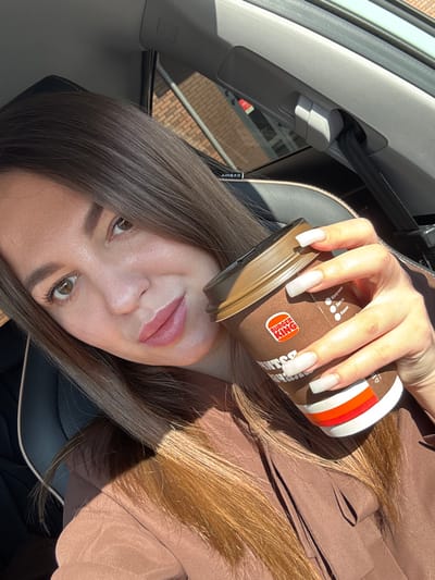 The scene is an eye-level, close-up photograph of a female subject seated inside a vehicle during daylight hours, contextualized to Likino-Dulyovo, Russia. The woman, who appears to be taking a selfie, occupies the left and central portions of the frame, looking directly towards the viewer with a slight smile. She has long, straight brown hair, brown eyes, and light-colored lip makeup. She is wearing a brown or taupe-colored garment, possibly a blouse or jacket, with visible folds and seams. Her left hand is prominently displayed, holding a disposable coffee cup. Her nails are long, squared, and light-colored, possibly white or very light nude.

The coffee cup is brown with a darker brown lid and features the Burger King logo. Partial Russian text, including "Капучино," "Латте," and "Кофе" along with what appears to be size indicators or prices, is visible on the cup. Below the text, there are horizontal stripes in white, orange, and red.

The interior of the vehicle is visible, featuring a black leather seat with a light-colored, possibly white or cream, trim line. Gray plastic panels form the vehicle's interior roof and pillar structures. An "AIRBAG" label is present on a panel near the upper right of the frame, and a seatbelt mechanism is visible on the B-pillar. Bright sunlight illuminates the scene from the right, casting shadows on the woman's face, hair, and the cup. Through the vehicle's window, a building constructed of reddish-brown bricks is visible in the background, along with a hint of a metal structure or railing.