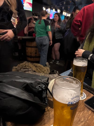 A crowded bar scene includes people standing and sitting. Two glasses of Birrificio Angelo Poretti beer sit on a wooden table, near a black leather bag and a brown furry item. Other people can be seen in the background, and a wooden barrel is visible near the center. The lighting is ambient, suggesting an indoor location.