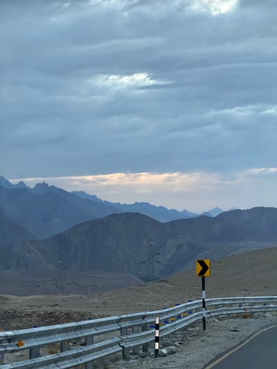 A scene along a winding mountain road in Chumkar, India, features a guardrail on the right side of the asphalt road. The guardrail, made of reflective metal, is adorned with blue and yellow markers. A yellow directional arrow sign with a black chevron points to the right. Beyond the road, a barren, rocky landscape stretches towards a series of imposing, layered mountains under a cloudy, overcast sky. The sky has a pale strip of light near the horizon, suggesting either sunrise or sunset. In the middle ground, several transmission towers stand against the backdrop of the mountains, with a few silhouetted birds visible around one of them. The overall impression is one of a remote, high-altitude environment.