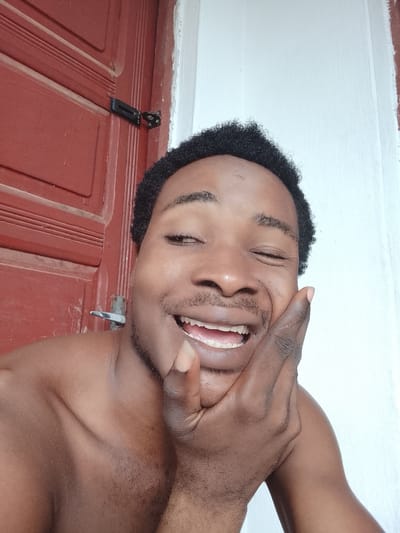 A close-up selfie captures a young Black man with curly hair, bare-chested, leaning against a red wooden door. His right hand is placed on his cheek, and he is smiling with his mouth open, his eyes partially closed. The background consists of the red door on the left and a plain white wall on the right. A silver door latch is visible on the red door. The scene appears to be outdoors, possibly on a porch or near an entrance in Orodo, Nigeria.